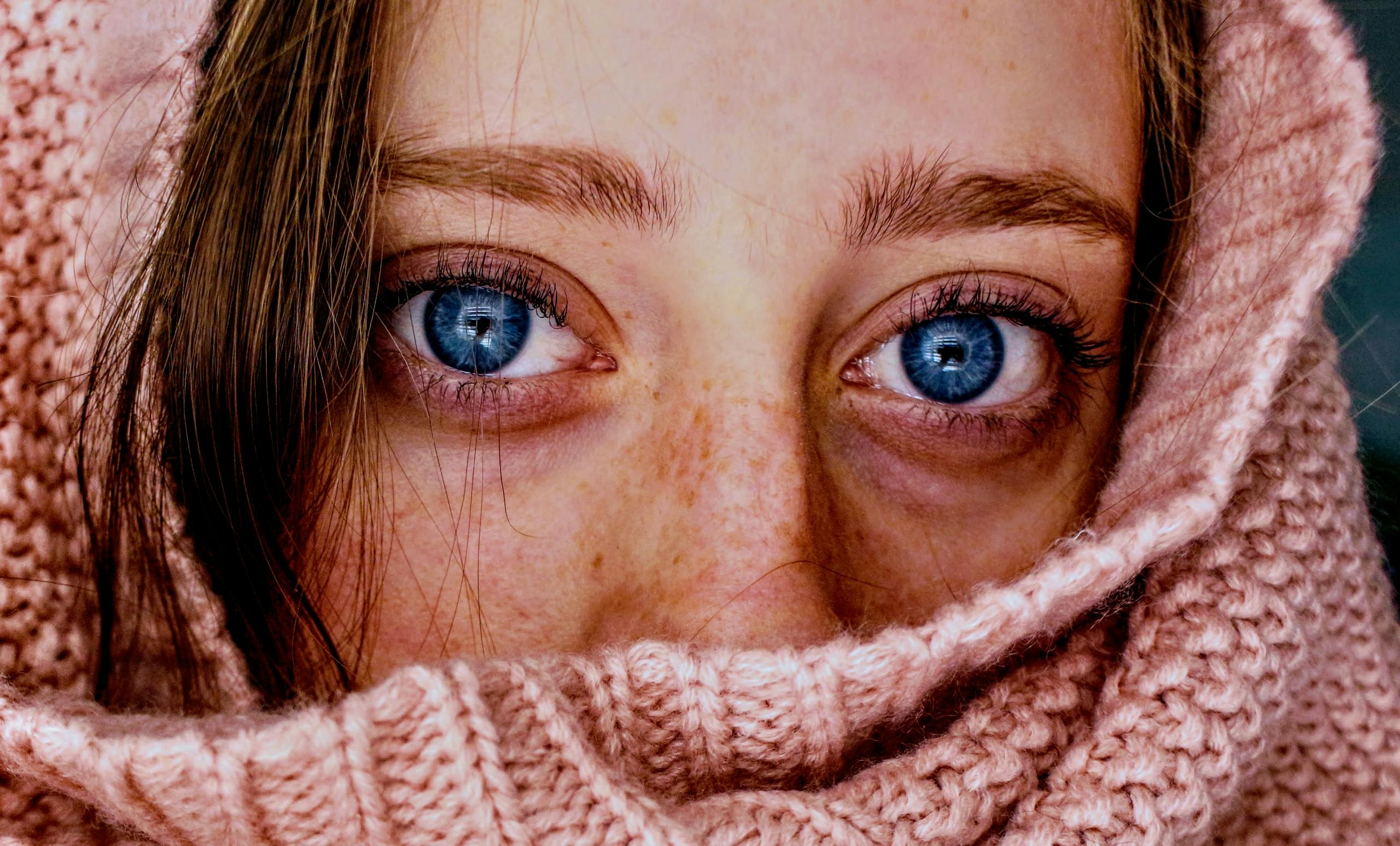 Intense close-up of a woman's blue eyes wrapped in a soft, pink knit scarf.