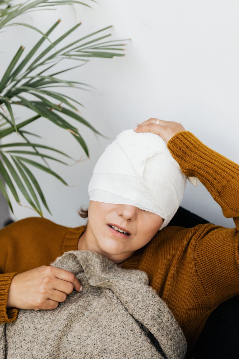 A woman with a bandaged head rests indoors, indicating a recent injury and seeking comfort.