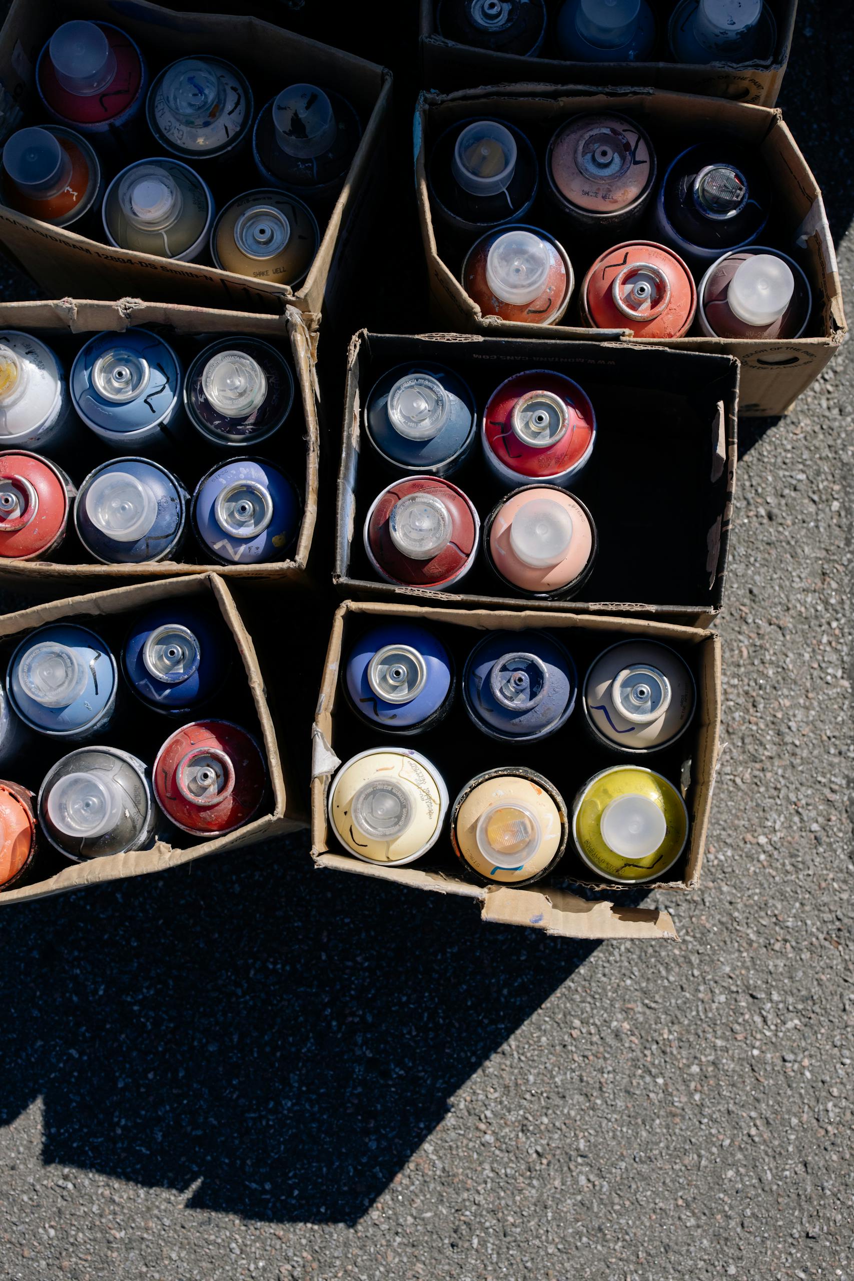 Top view of various spray paint cans organized in cardboard boxes on pavement.
