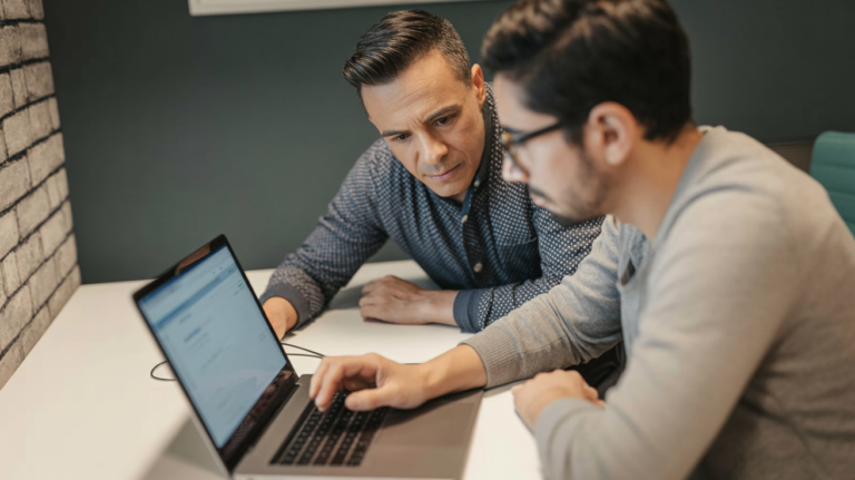Manager leaning over a developer's shoulder, both focused on a laptop chat interface — conveying trust and scrutiny.