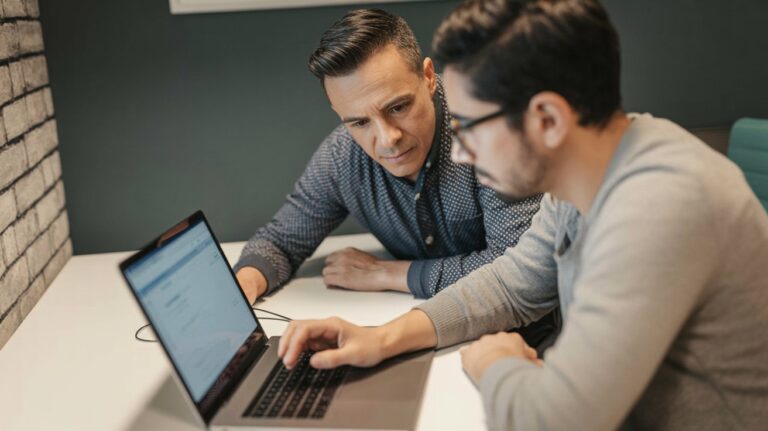 Manager leaning over a developer's shoulder, both focused on a laptop chat interface &mdash; conveying trust and scrutiny.